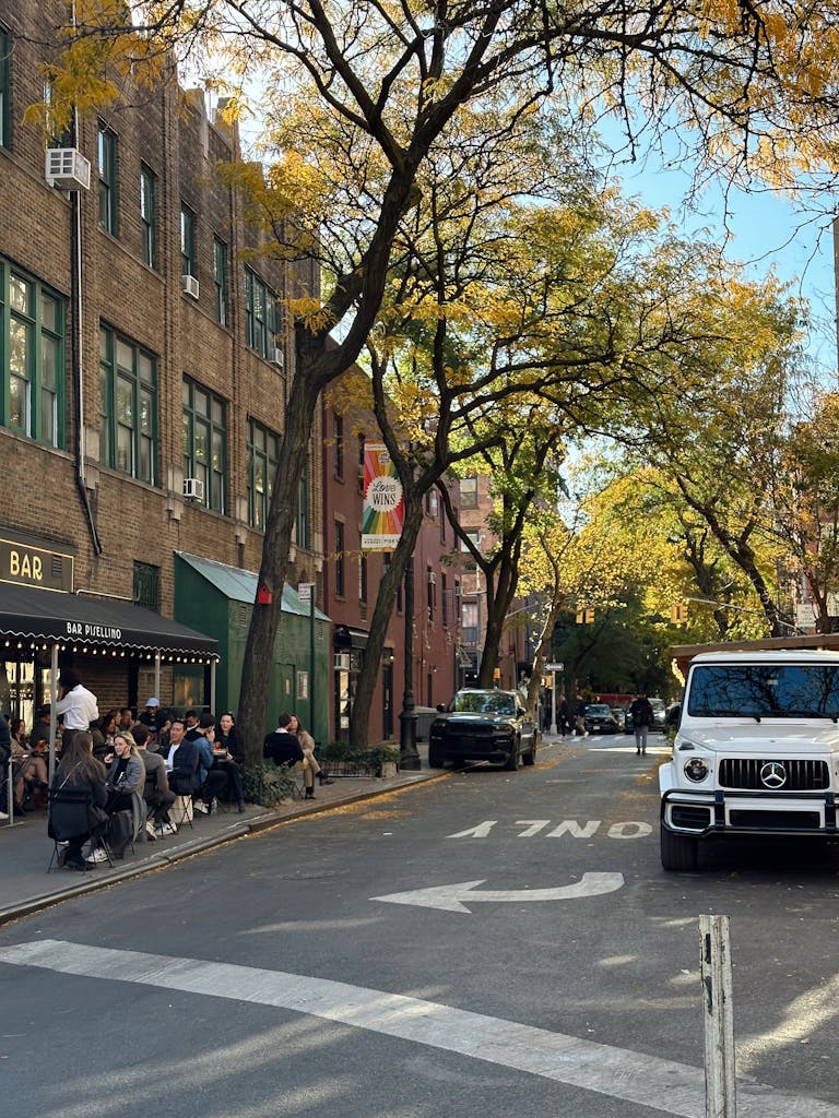 Autumn city street with outdoor seating, trees, and parked cars under warm light.