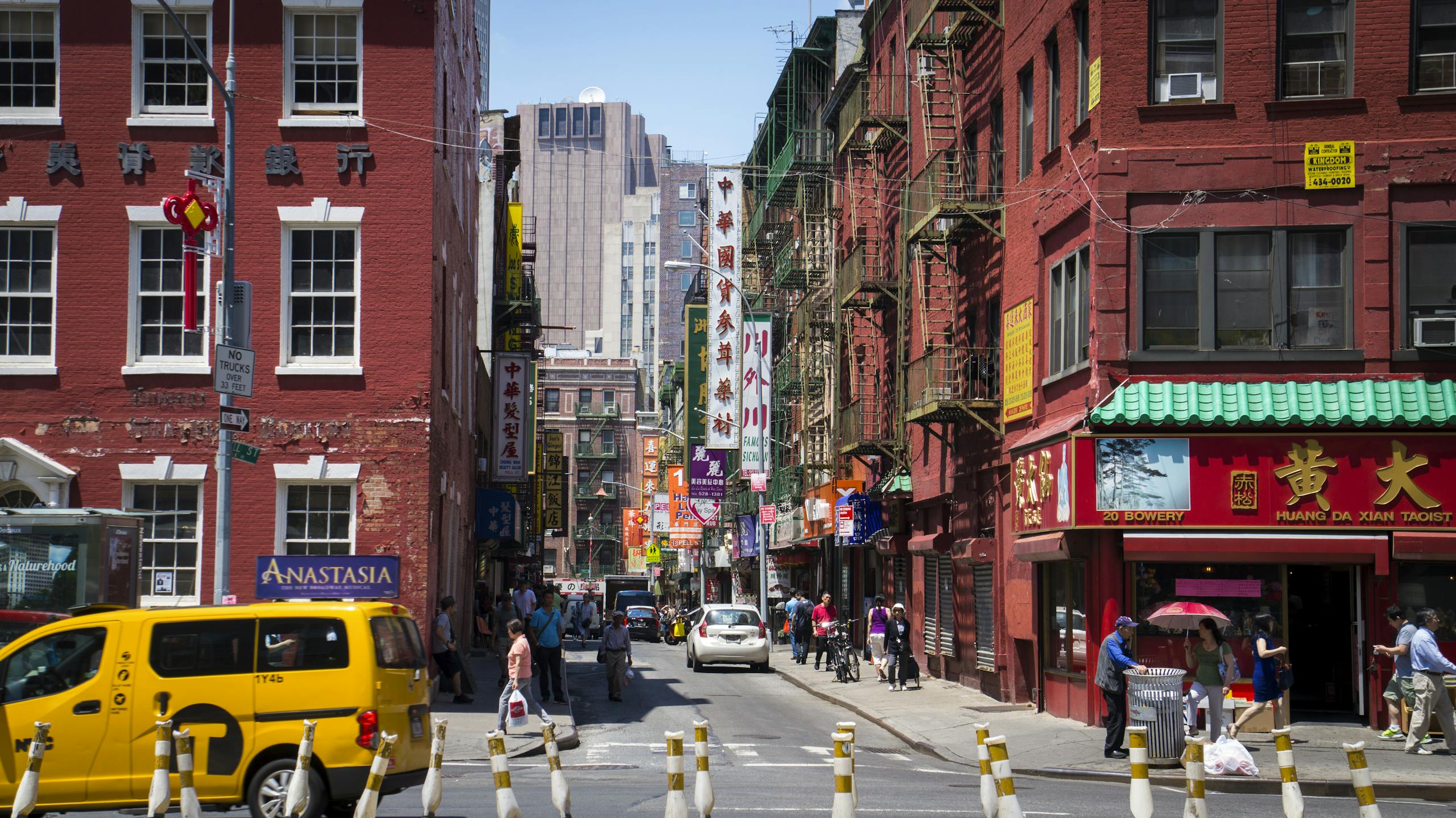 Busy daytime street scene in Chinatown, New York City with pedestrians, shops, and taxis.
