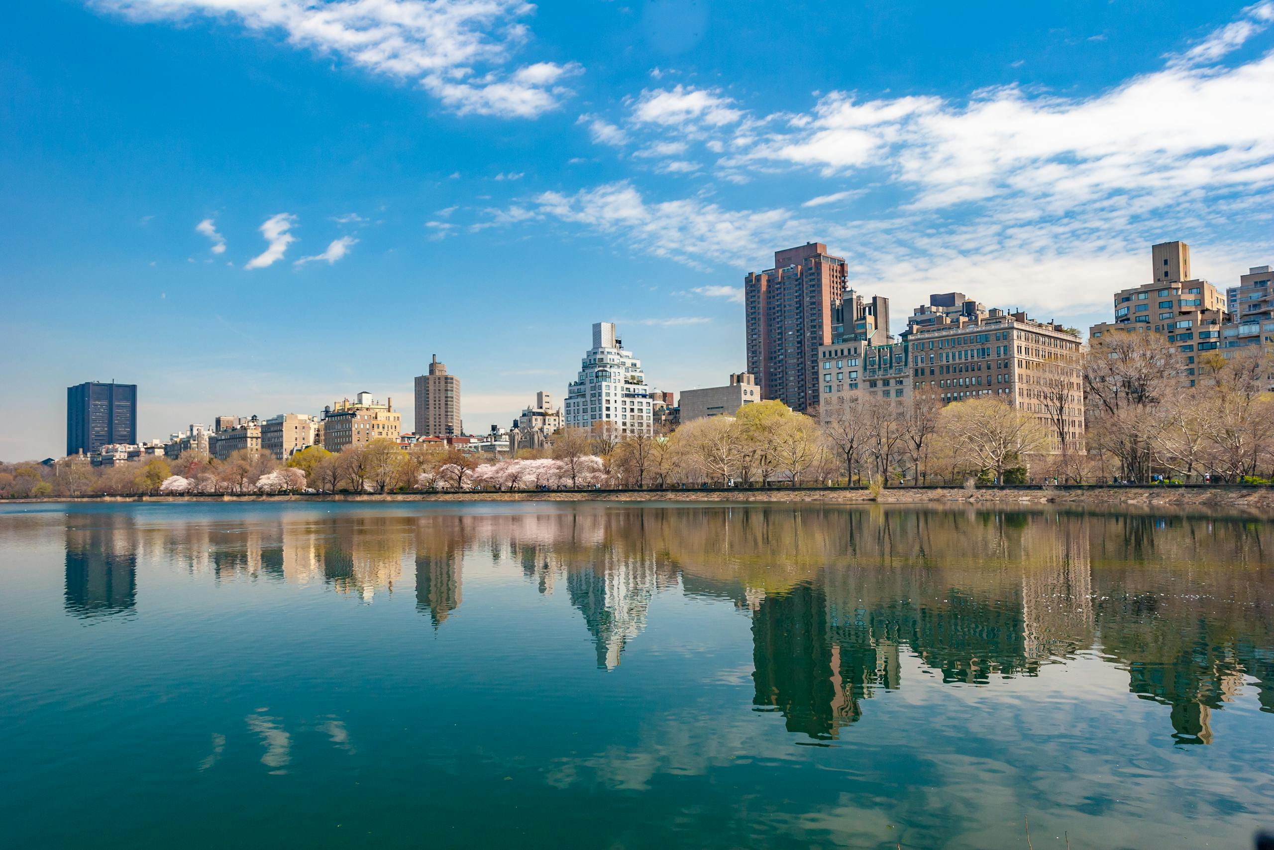 New York City skyline reflecting in Central Park's serene water under a vibrant blue sky.